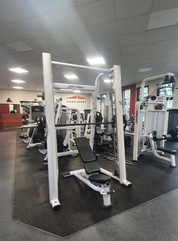 Ein moderner Fitnessraum mit weißen Geräten und gepolsterten schwarzen Sitzen, im Hintergrund trainieren Personen.