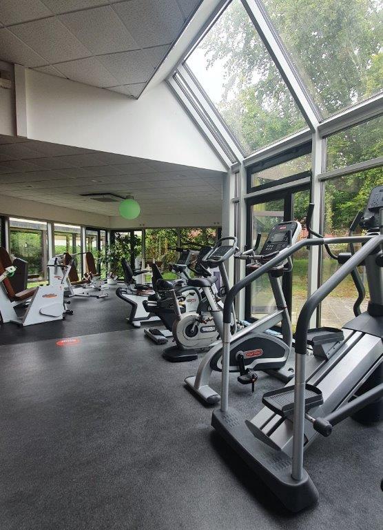 Fitnessstudio mit leeren modernen Geräten, belichtet durch große Glasfenster, Blick ins Grüne.