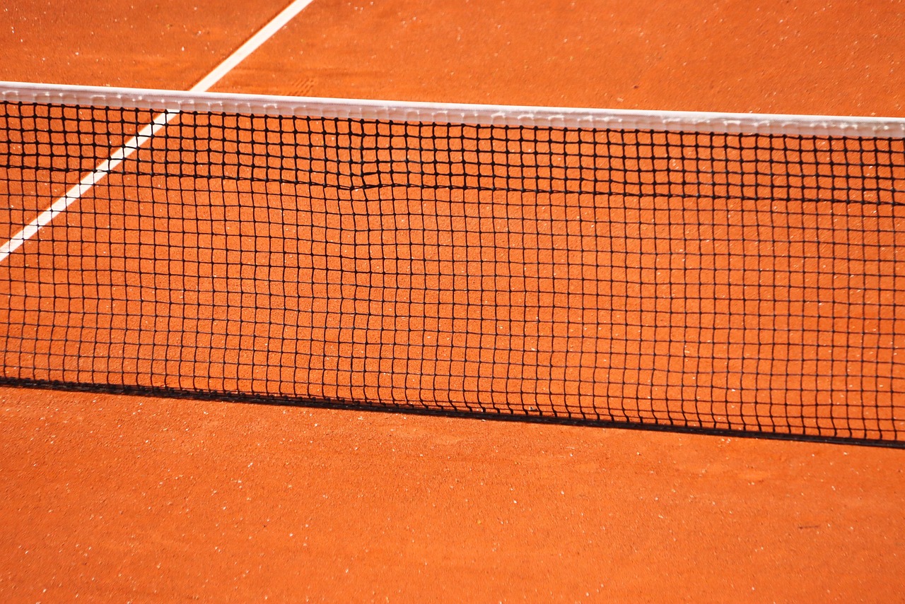 Tennisplatz mit orangefarbenem Sandboden und Netz in der Mitte, diagonale Linien im Hintergrund.